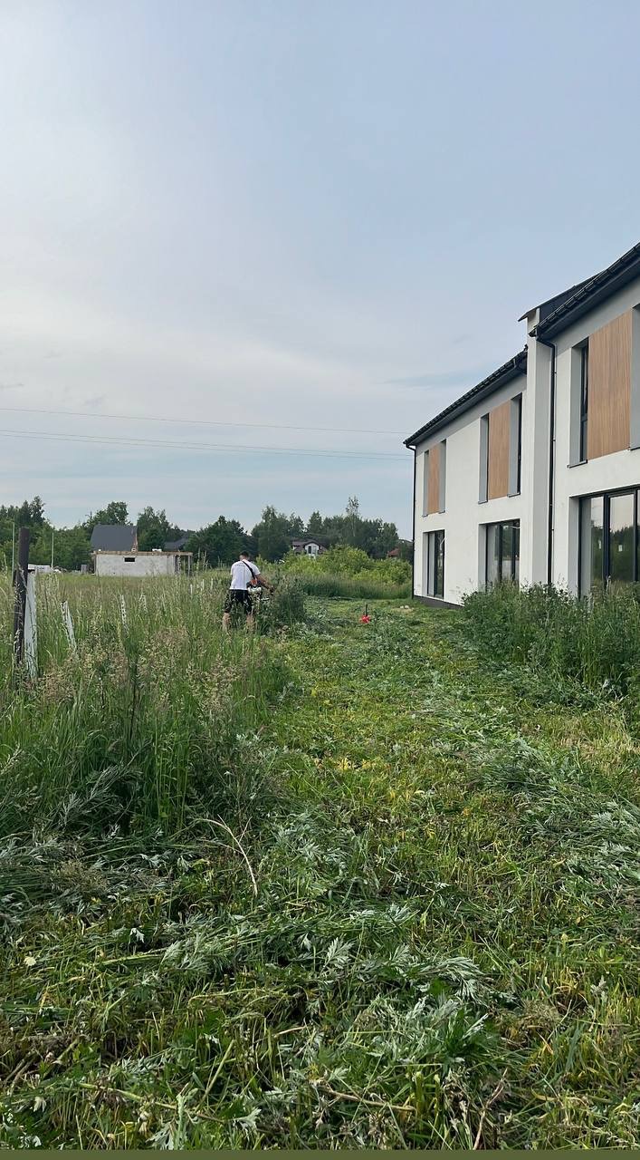 Mężczyzna kosi wysoką trawę przy nowoczesnym domu. Świeżo skoszona trawa leży na ziemi, ukazując postęp pracy. Tło: budynki i zieleń.