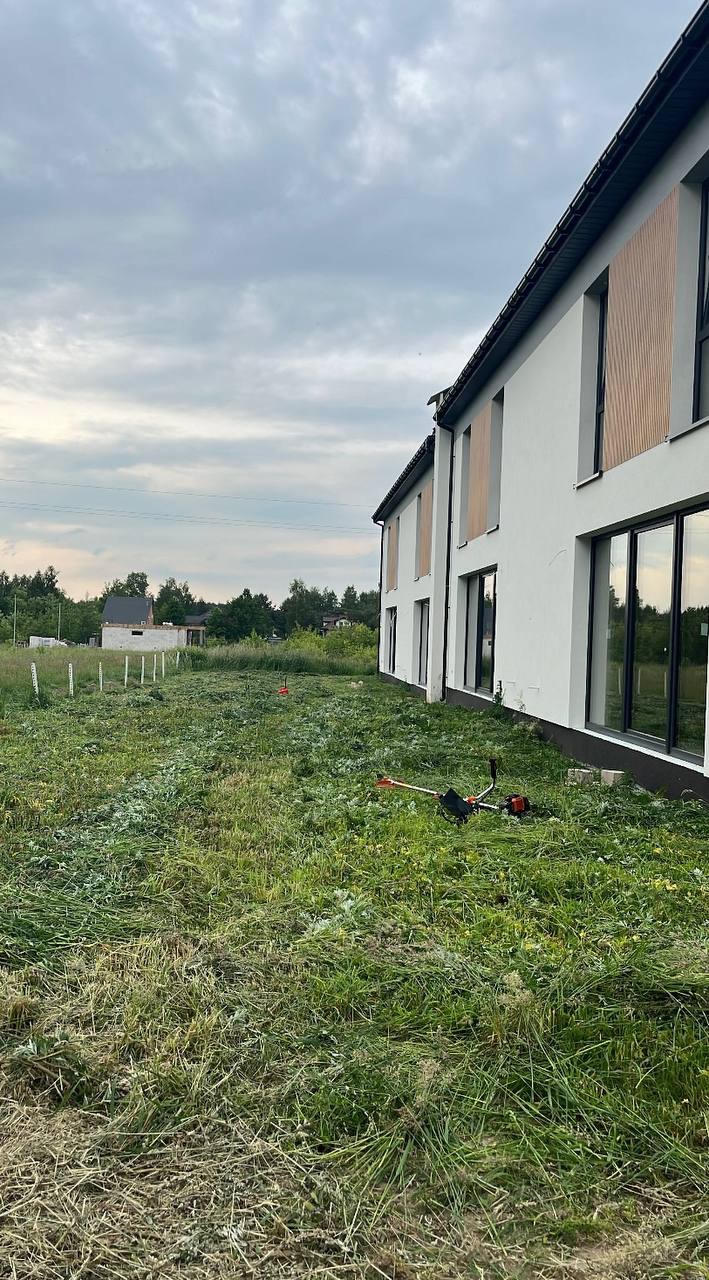 Częściowo skoszona trawa na działce obok nowoczesnego domu z dużymi oknami. Na trawie leży kosa spalinowa. W tle widoczne drzewa i budynek w budowie.