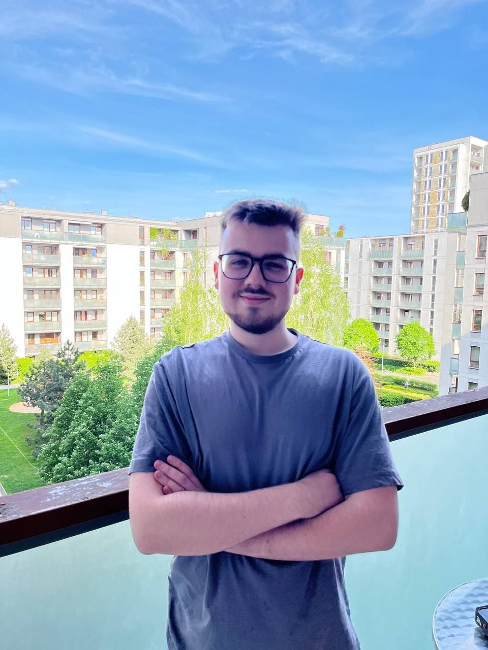 Młody mężczyzna w okularach, stojący na balkonie z założonymi rękami, na tle miejskiego krajobrazu z budynkami i zielenią w słoneczny dzień.