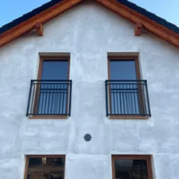 Elewacja budynku z oknami w brązowych ramach i czarnymi, metalowymi balustradami balkonowymi. Widok z dołu na szczyt budynku pod błękitnym niebem.