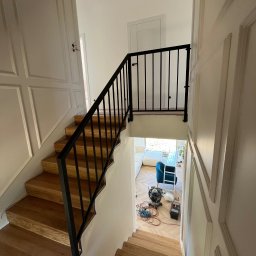 ATU-SPAW - Drewniane schody z czarną, metalową balustradą w jasnym wnętrzu. Widok z góry, na dole widoczne narzędzia i zabezpieczenia podłogi, co sugeruje prace wykończeniowe.