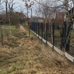 ATU-SPAW - Ciemne ogrodzenie panelowe z podmurówką betonową na nierównym terenie porośniętym suchą trawą, widoczne drzewa bez liści i fragment budynku w tle.