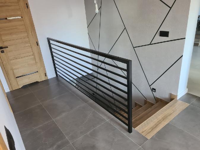 Nowoczesna, czarna balustrada schodowa z poziomych profili, na tle ściany z geometrycznym wzorem i drewnianych schodów. Minimalistyczny design, jasne wnętrze.