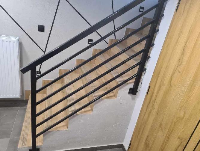 Nowoczesna, czarna balustrada metalowa z poziomymi szczeblami na drewnianych schodach, minimalistyczny design, widok z dołu, jasne wnętrze z dekoracyjnymi liniami na ścianie.