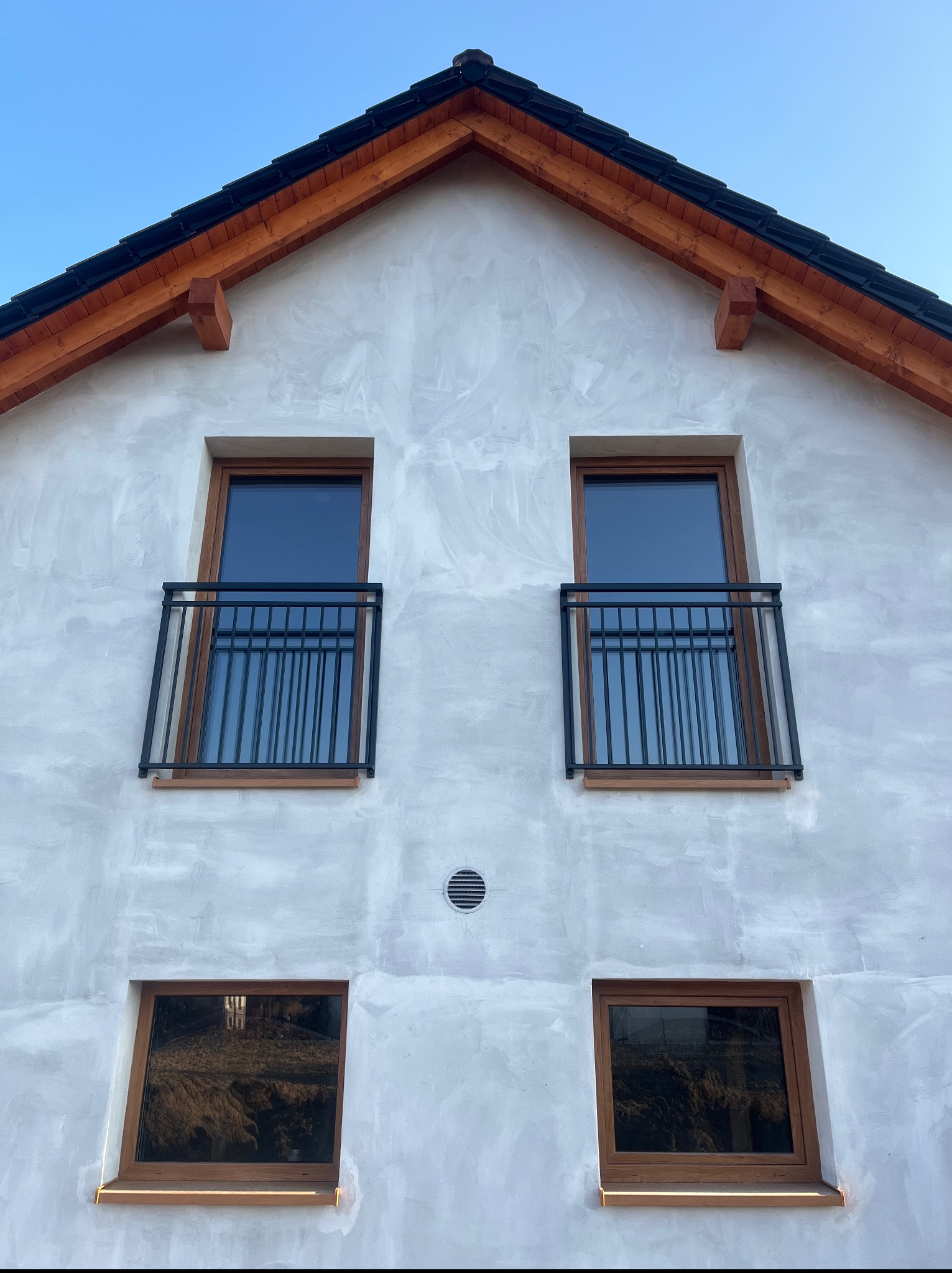 Elewacja budynku z oknami w brązowych ramach i czarnymi, metalowymi balustradami balkonowymi. Widok z dołu na szczyt budynku pod błękitnym niebem.