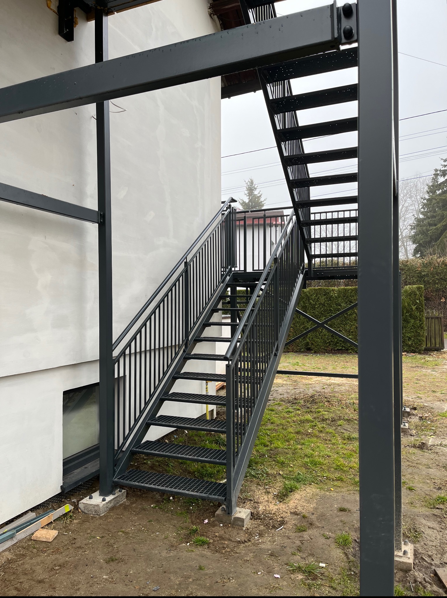 Zewnętrzne schody metalowe z poręczami, prowadzące na wyższą kondygnację budynku. Konstrukcja pomalowana na ciemny kolor, widoczne elementy montażowe.