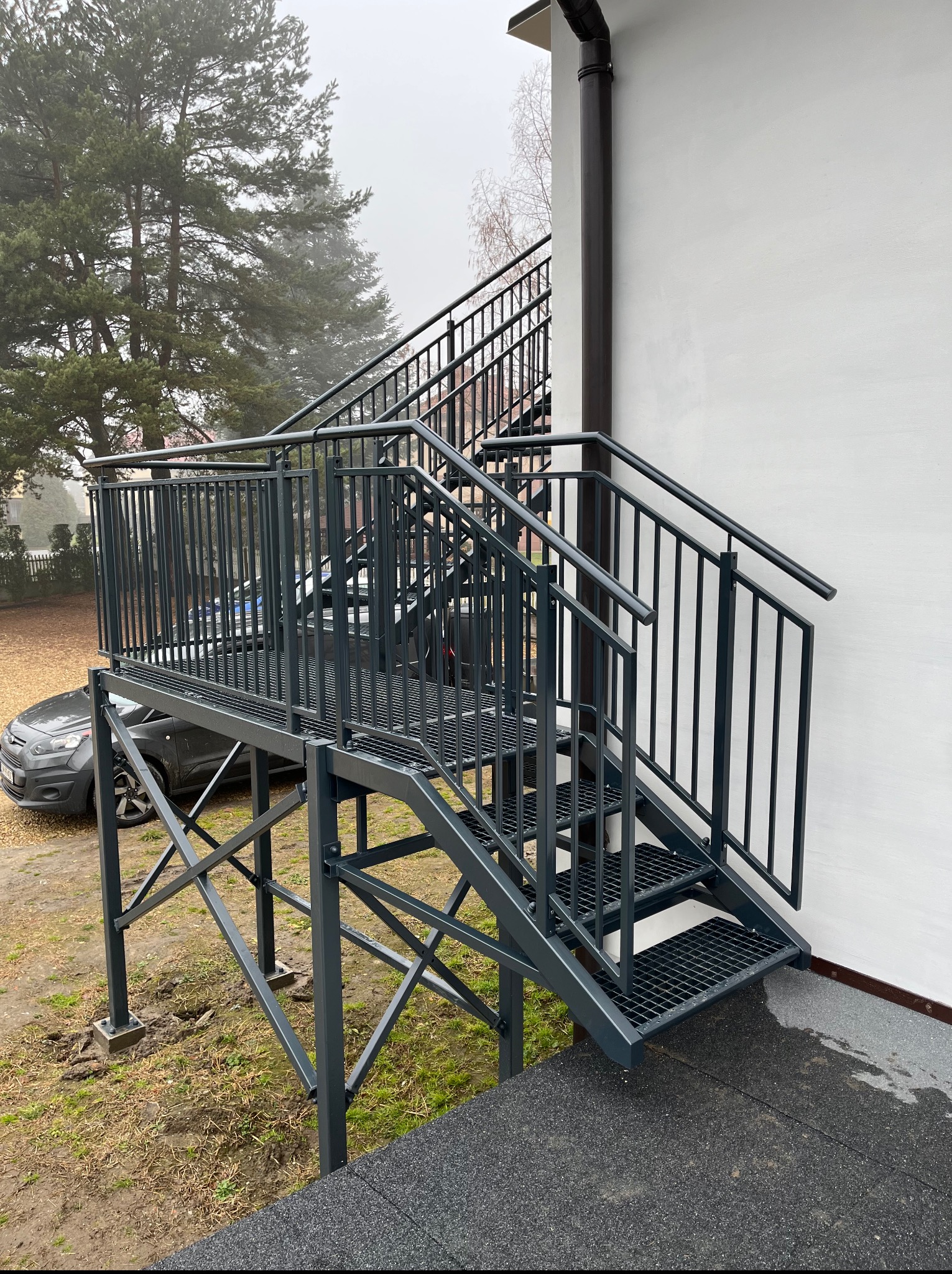 Zewnętrzne schody metalowe z poręczami, prowadzące do budynku z białą elewacją. Konstrukcja pomalowana na szaro, widoczny samochód w tle. Ujęcie z dołu, pochmurny dzień.