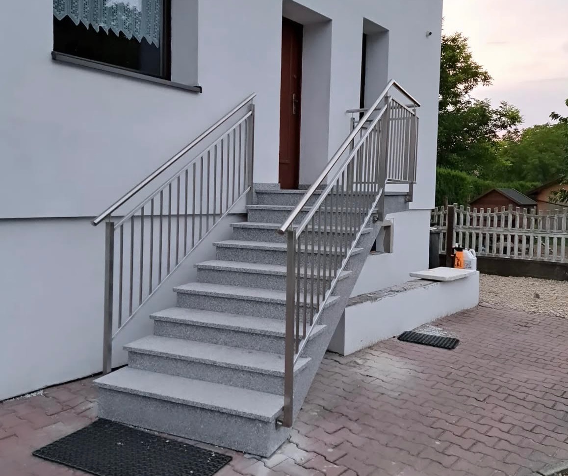 Zewnętrzne schody granitowe z nowoczesną balustradą ze stali nierdzewnej, prowadzące do drzwi wejściowych domu. Widok z dołu, ceglana kostka brukowa.