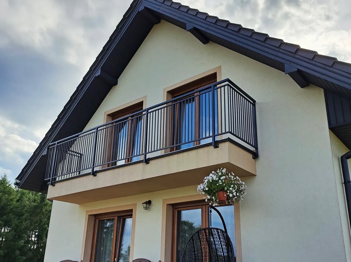 Elewacja domu z balkonem i czarną, metalową balustradą. Na balkonie widoczne okna z drewnianą ramą. Pod balkonem wisząca donica z białymi kwiatami.