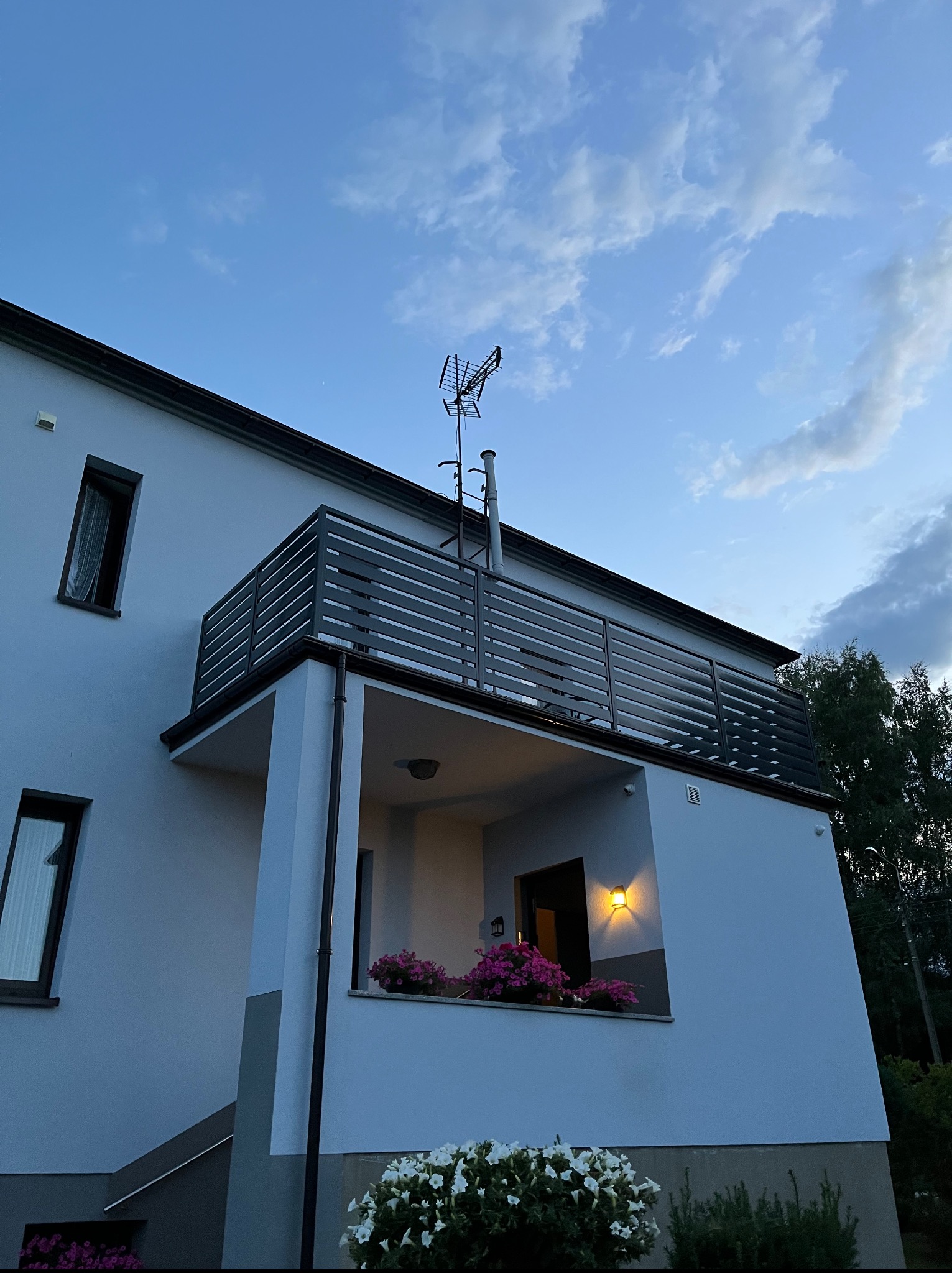 Nowoczesny balkon z szarą, poziomą balustradą na tle błękitnego nieba. Kwiaty w donicach dodają uroku. Widok z dołu podkreśla architekturę budynku.