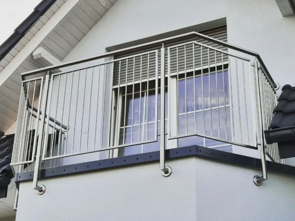 Balkon z nowoczesną, metalową balustradą ze stali nierdzewnej. Pionowe pręty, gładka powierzchnia, widoczne mocowania. Tło: biała elewacja budynku i okno z żaluzjami.