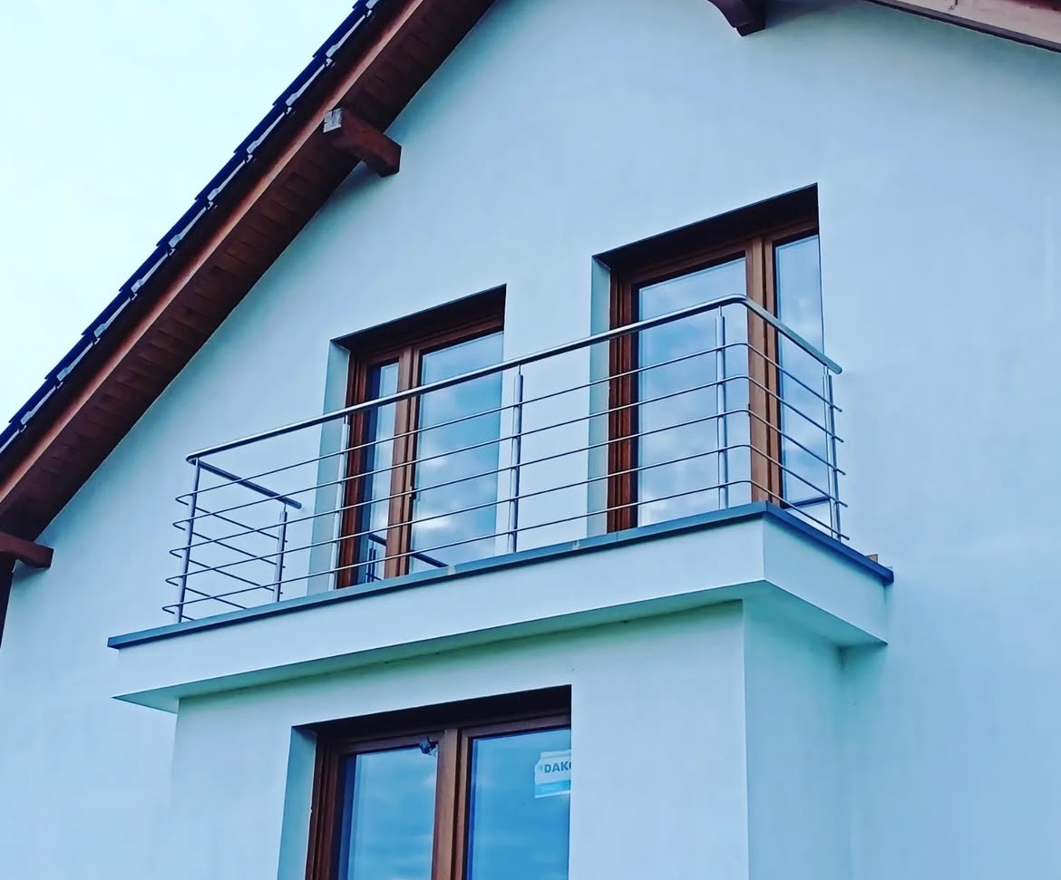Balkon z nowoczesną, minimalistyczną balustradą ze stali nierdzewnej, widok z dołu, na tle jasnej elewacji domu z drewnianymi elementami dachu i ramami okiennymi.