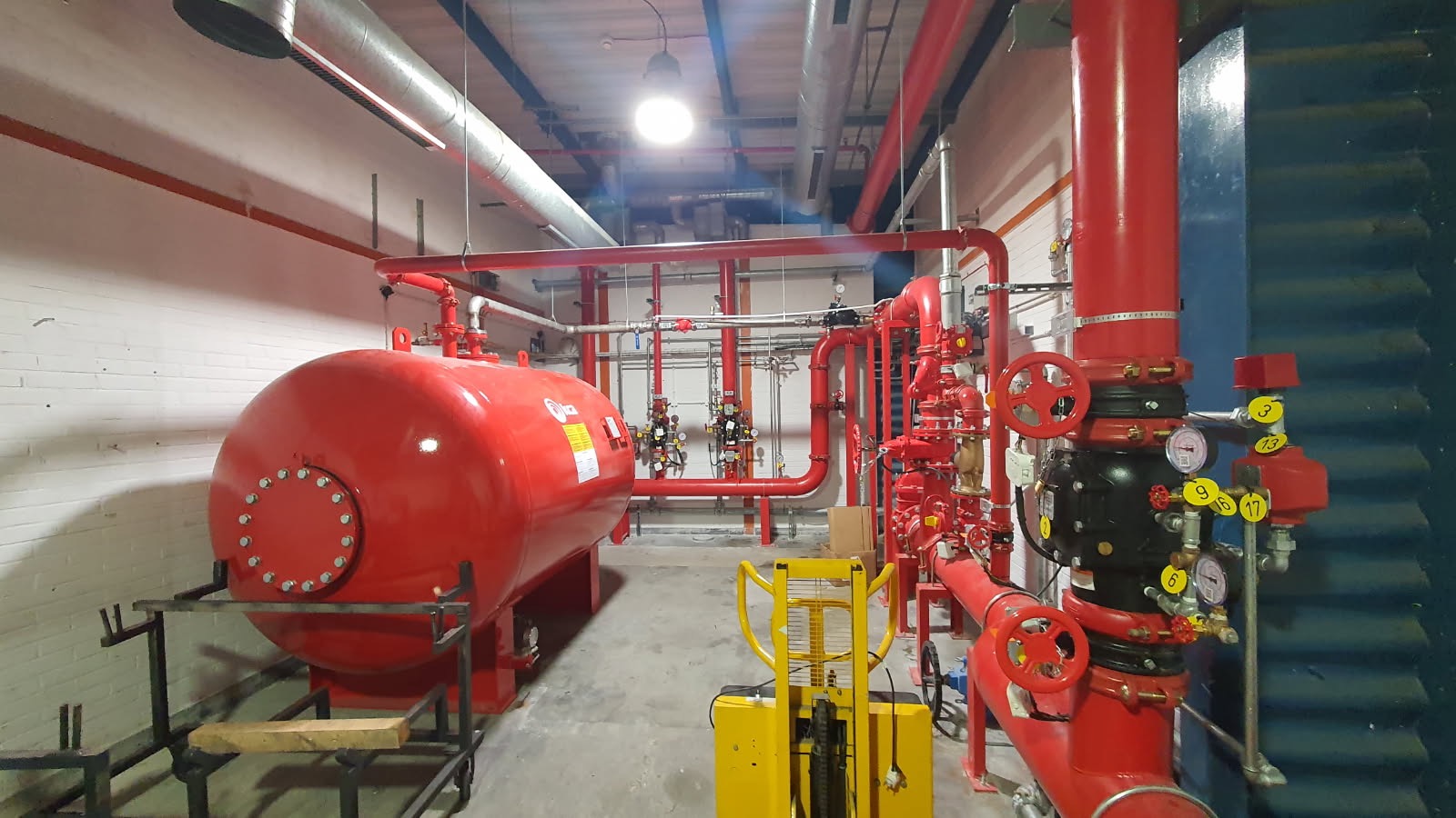 Kompleksowa instalacja przeciwpożarowa z czerwonym zbiornikiem ciśnieniowym i rurami, widoczna w hali przemysłowej. Elementy instalacji oznaczone numerami.