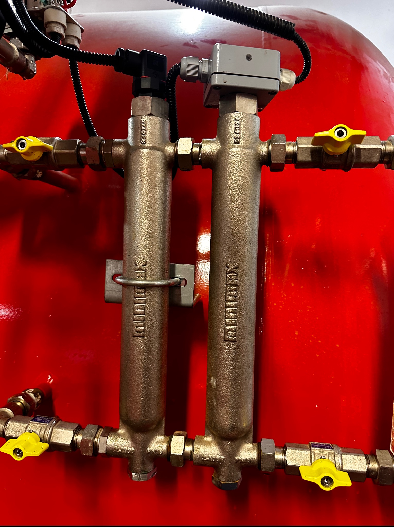 Czerwony zbiornik z dwoma mosiężnymi rozdzielaczami Minimax, z żółtymi zaworami, podłączonymi przewodami i czujnikiem. Element instalacji grzewczej.