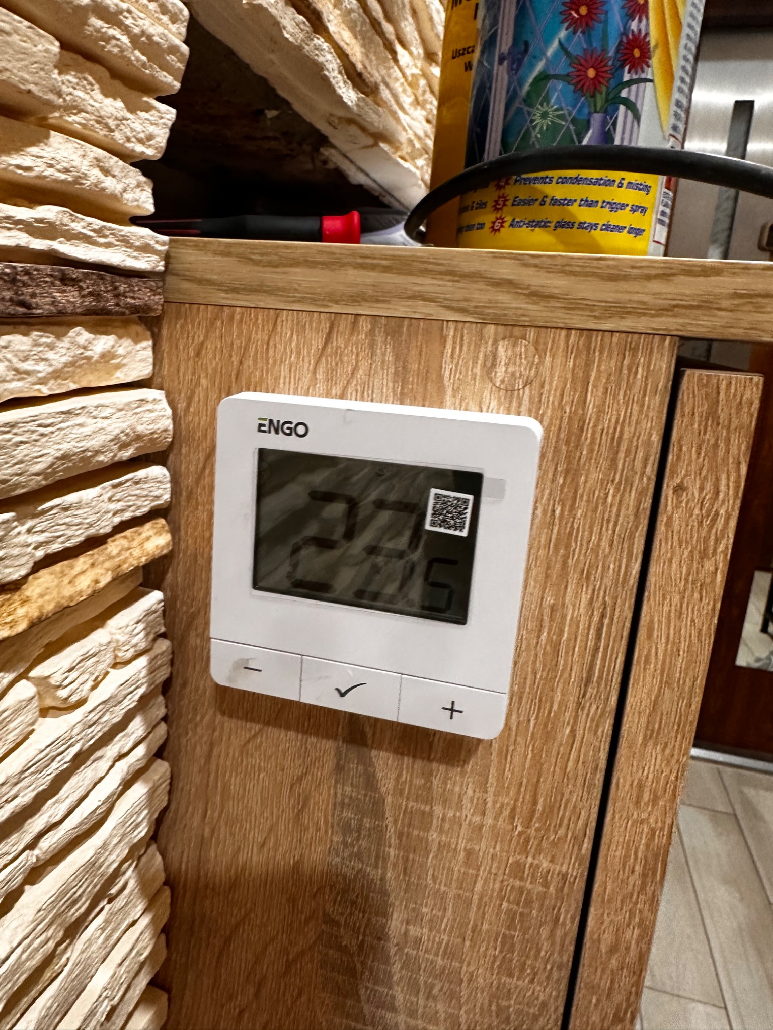 Panel sterowania ogrzewaniem ENGO, zamontowany na drewnianej szafce obok dekoracyjnej ściany z kamienia. Widoczny wyświetlacz z temperaturą i przyciski regulacji.