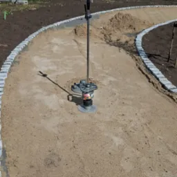 Ubijarka wibrostopowa zagęszcza piasek na ścieżce z obrzeżami z kostki granitowej. W tle budynek Kampex i zaparkowane pojazdy.