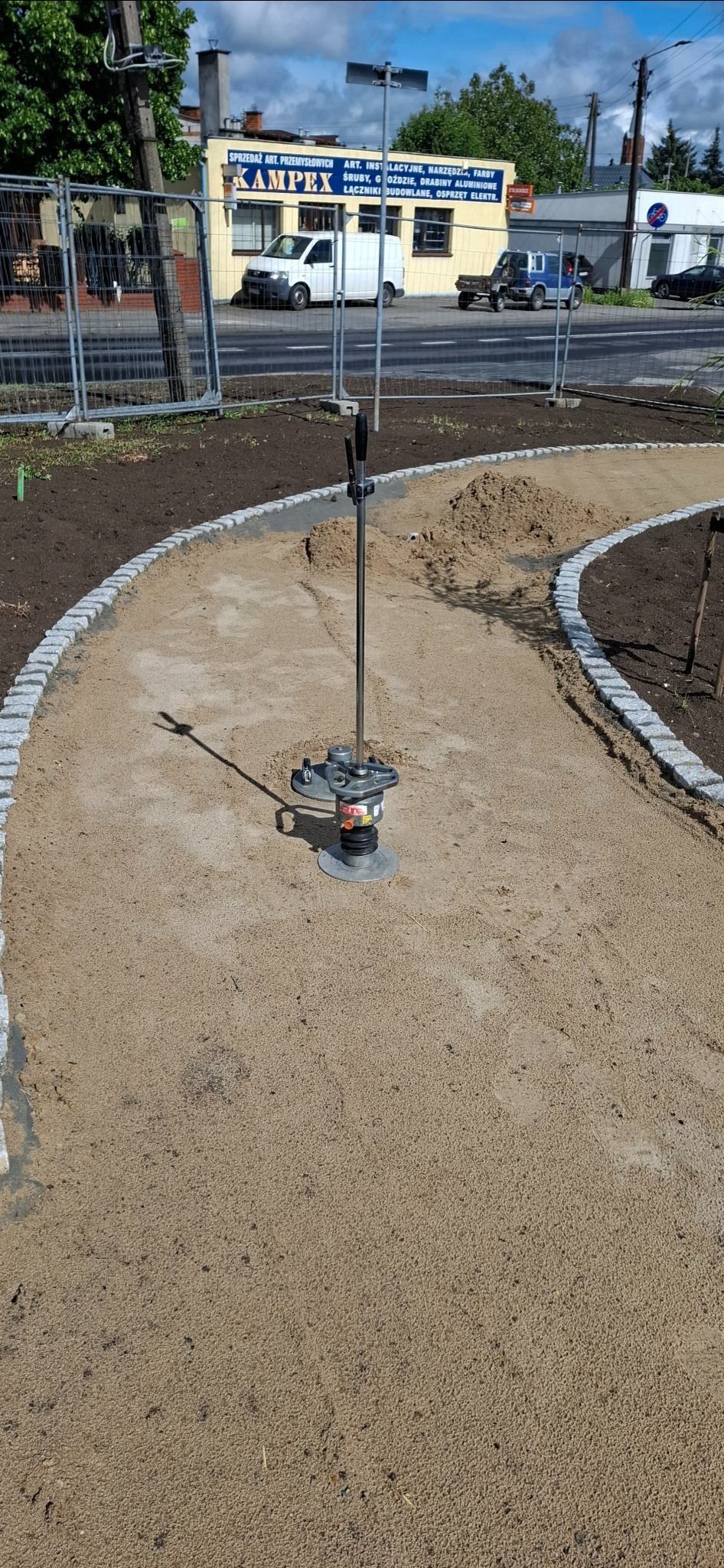 Ubijarka wibrostopowa zagęszcza piasek na ścieżce z obrzeżami z kostki granitowej. W tle budynek Kampex i zaparkowane pojazdy.