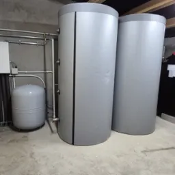 Kompletna instalacja pompy ciepła w kotłowni: dwa szare zbiorniki akumulacyjne, zasobnik, rury i czerwony piec na pellet. Widok na urządzenia w surowym pomieszczeniu.