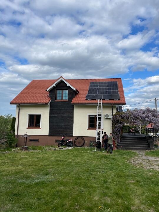 Montaż paneli fotowoltaicznych na dachu domu jednorodzinnego z czerwoną dachówką, przy użyciu drabiny i w obecności dwóch monterów. Błękitne niebo w tle.