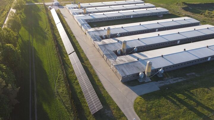 Widok z góry na farmę z panelami fotowoltaicznymi obok budynków. Panele ciągną się wzdłuż drogi, a budynki mają szare dachy i białe ściany.