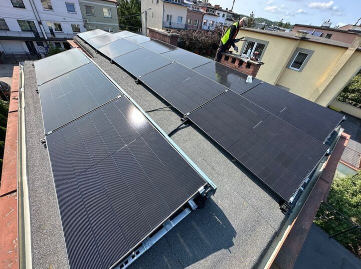 Panele fotowoltaiczne na dachu budynku mieszkalnego, montowane przez instalatora w odblaskowej kamizelce, w słoneczny dzień. Widok z góry.