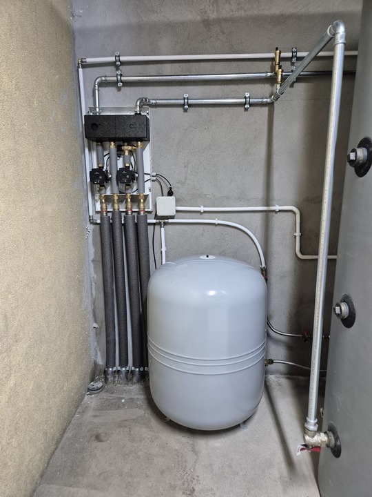 Szara instalacja z rurami, zbiornikiem wyrównawczym i rozdzielaczem, zamontowana na surowej, betonowej ścianie w pomieszczeniu technicznym. Elementy instalacji zabezpieczone.