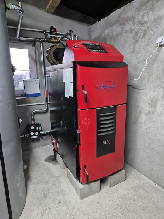 Czerwono-czarny piec Attack SLX, ustawiony na betonowych bloczkach w surowym pomieszczeniu. Widoczne rury instalacji, szary zbiornik i gniazdko elektryczne.