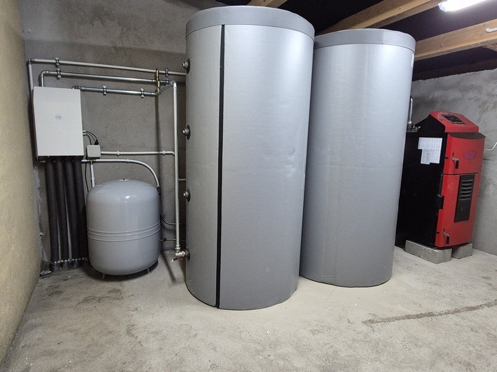 Kompletna instalacja pompy ciepła w kotłowni: dwa szare zbiorniki akumulacyjne, zasobnik, rury i czerwony piec na pellet. Widok na urządzenia w surowym pomieszczeniu.