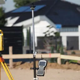 Żółty teodolit na statywie i tyczka geodezyjna z kontrolerem Trimble w terenie budowy, w tle dom z panelami słonecznymi.