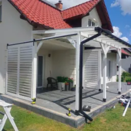 Biała pergola z przesuwnymi panelami żaluzjowymi, częściowo przysłaniająca taras. W tle dom z czerwoną dach&oacute;wką i narzędzia pracy na trawniku.