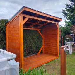 HOLZMET - Brązowa, otwarta altana z drewna, z czarnym dachem, na trawniku. W tle zielone krzewy i drzewa. Widoczne elementy montażowe i konstrukcyjne.