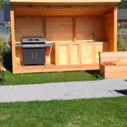 Drewniana altana z grillem i zlewem na trawniku, oświetlona sznurem lampek. Widoczny fragment ławki. Słoneczny dzień, błękitne niebo w tle.