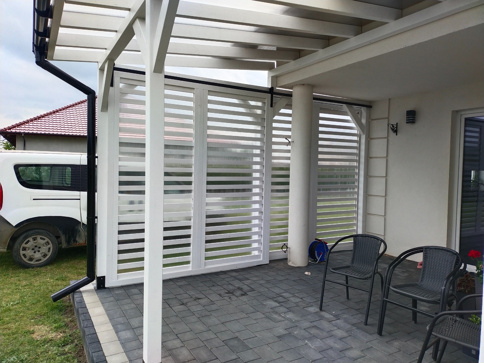 Nowoczesna zabudowa balkonu z białymi, przesuwnymi panelami żaluzjowymi, zapewniająca prywatność i cień. Widoczne krzesła i kostka brukowa na tarasie.