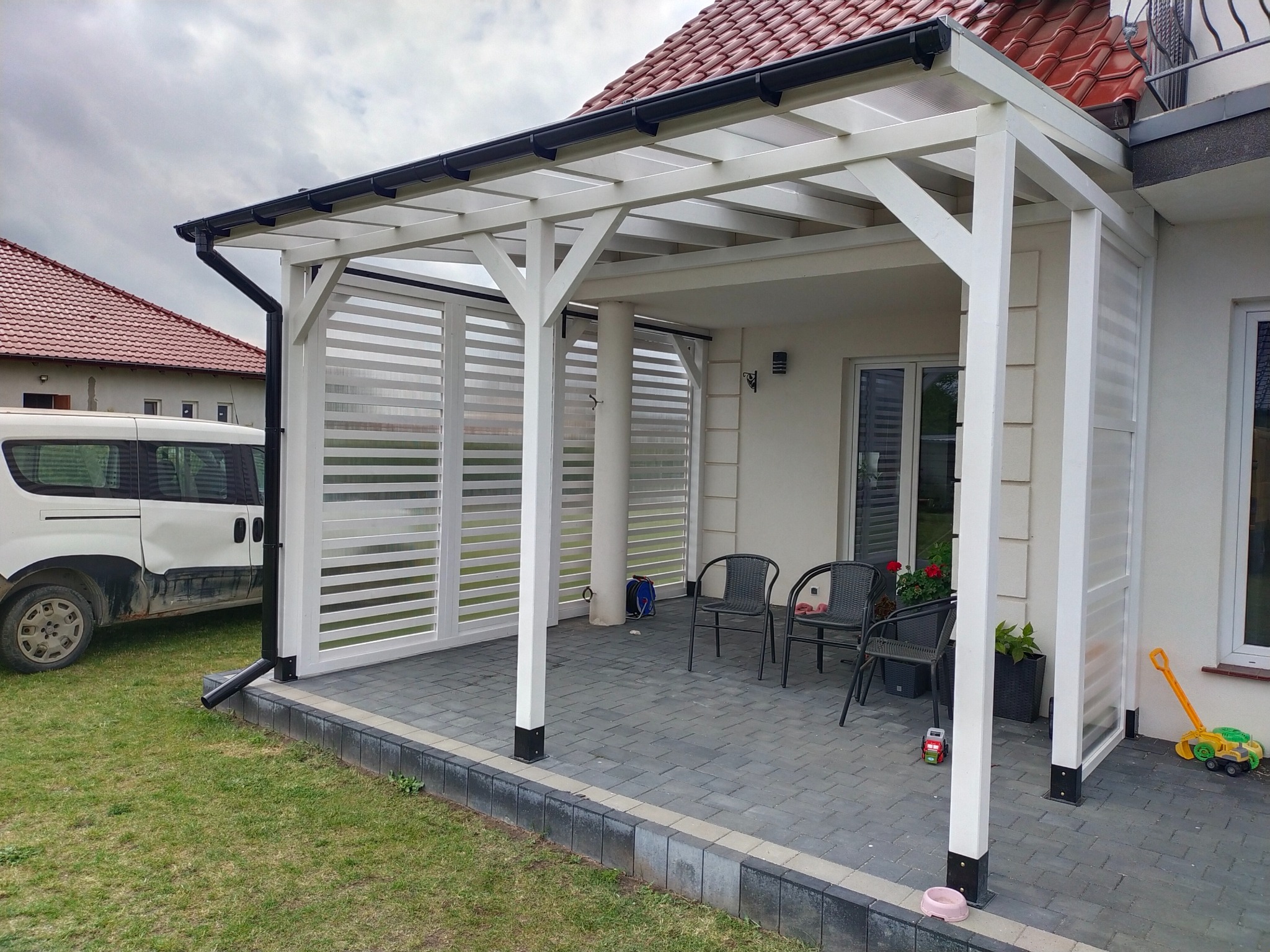 Biała pergola z osłonami przeciwsłonecznymi, zadaszona kostką brukową, z czarnymi rynnami i elementami wykończenia. Widoczny fragment elewacji domu i trawnik.