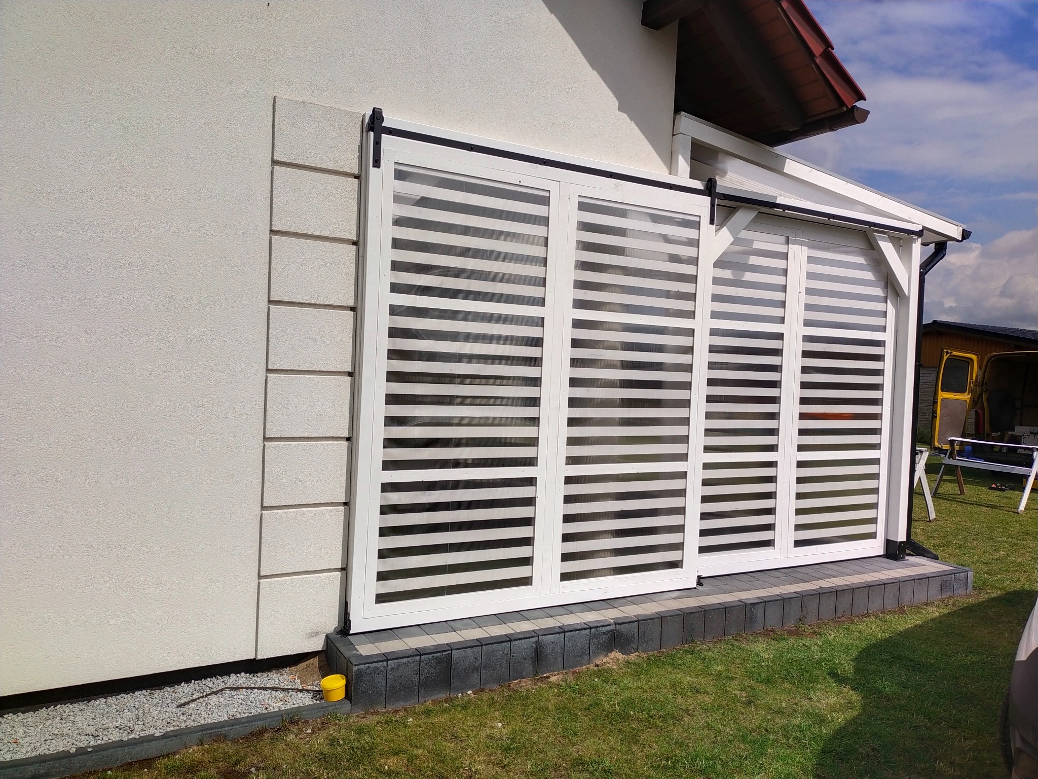 Nowoczesna zabudowa balkonu z przesuwnymi panelami żaluzjowymi w białej ramie, widoczna z zewnątrz, na tle trawnika i fragmentu budynku. Czarny system przesuwny.