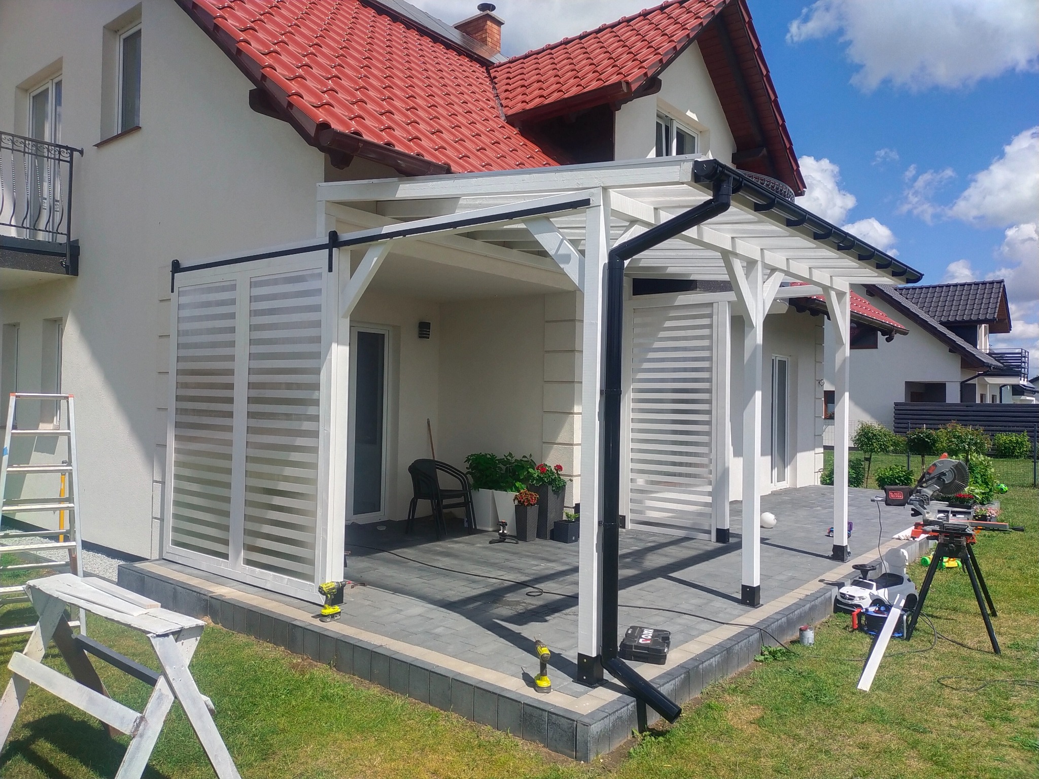 Biała pergola z przesuwnymi panelami żaluzjowymi, częściowo przysłaniająca taras. W tle dom z czerwoną dachówką i narzędzia pracy na trawniku.