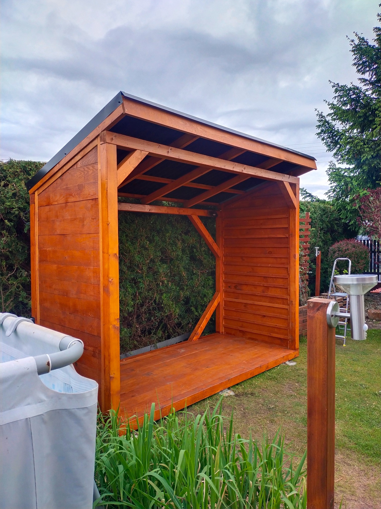 Brązowa, otwarta altana z drewna, z czarnym dachem, na trawniku. W tle zielone krzewy i drzewa. Widoczne elementy montażowe i konstrukcyjne.