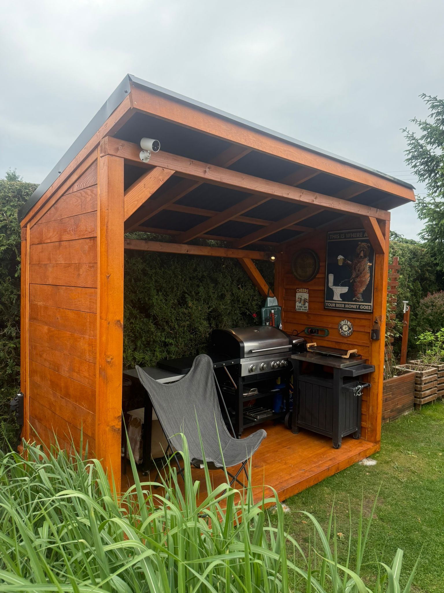 Drewniana altana z grillem i krzesłem na trawniku. Na ścianie plakaty i kamera monitoringu. Drewno w ciepłym, brązowym odcieniu. Otoczenie zielone, zarośnięte.