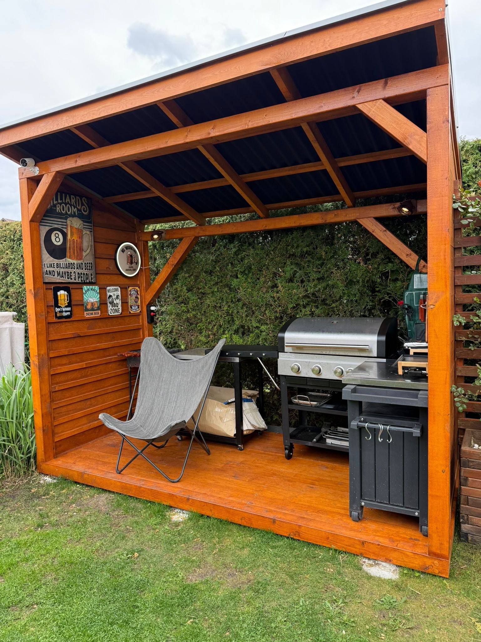 Drewniana altana z grillem i krzesłem na trawie. Wnętrze zdobią tematyczne plakaty. Widoczne oświetlenie i elementy dekoracyjne. Całość tworzy przytulną przestrzeń do grillowania.