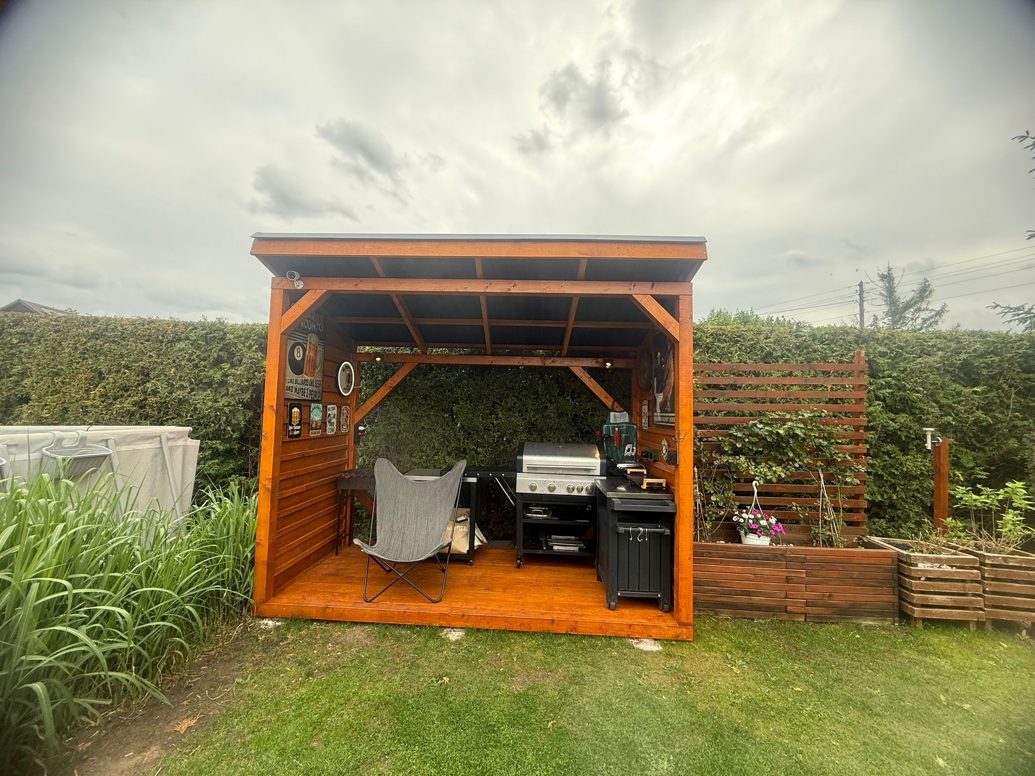 Drewniana altana z grillem i miejscem do siedzenia, otoczona zielenią. Widoczne dekoracje ścienne i rośliny w donicach. Całość w ogrodowej aranżacji pod pochmurnym niebem.