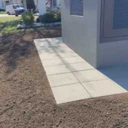 Ułożona ścieżka z jasnych płyt chodnikowych na świeżo przygotowanym gruncie, obok szarej budki technicznej z żaluzjami wentylacyjnymi.