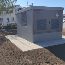 Rafał Sielski - Szara stacja transformatorowa NÖ Netz EVN na nowo przygotowanym terenie z kostką brukową, dom w tle i błękitne niebo. Widoczne ślady po pracach ziemnych.