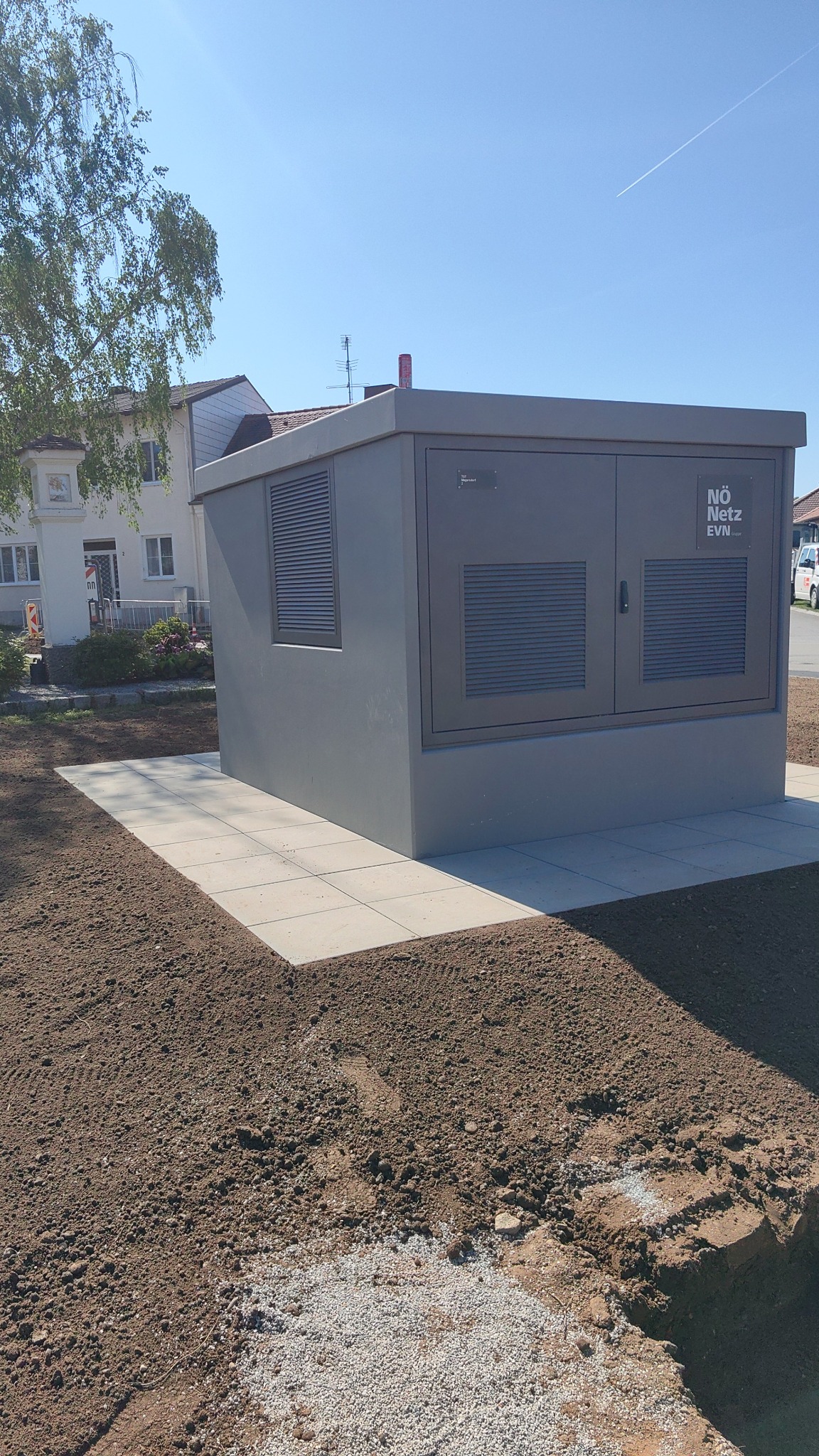 Szara stacja transformatorowa NÖ Netz EVN na nowo przygotowanym terenie z kostką brukową, dom w tle i błękitne niebo. Widoczne ślady po pracach ziemnych.