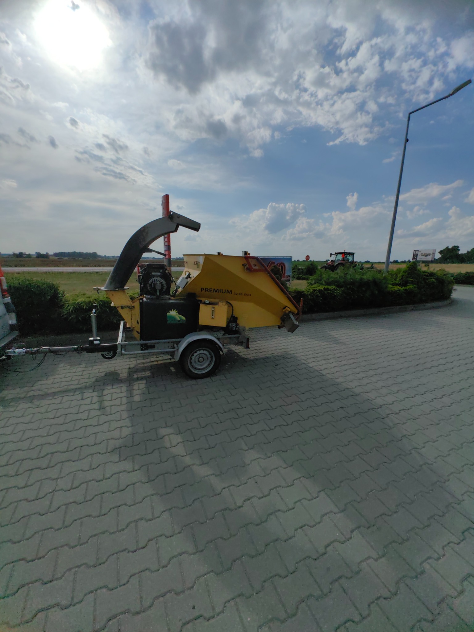 Żółto-czarna rębarka do gałęzi Premium 220 EVO na przyczepie, zaparkowana na kostce brukowej, z zielenią w tle i błękitnym niebem.