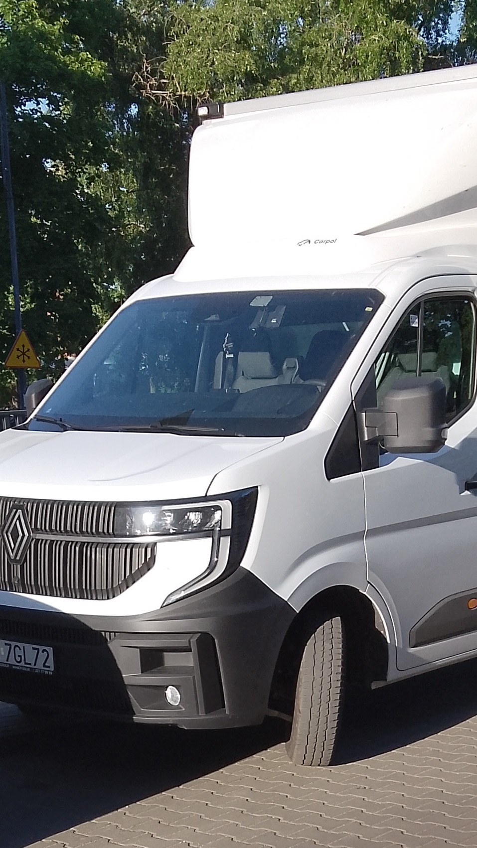 Biały samochód dostawczy Renault Master z wysoką zabudową na tle zieleni miejskiej, widoczny fragment tablicy rejestracyjnej i znak ostrzegawczy.