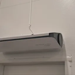 Biała klimatyzacja Flowair Slim zamontowana na ścianie z blachy trapezowej. Widoczna instalacja elektryczna i kamera przemysłowa. Tabliczka informacyjna 3.6m z lewej.