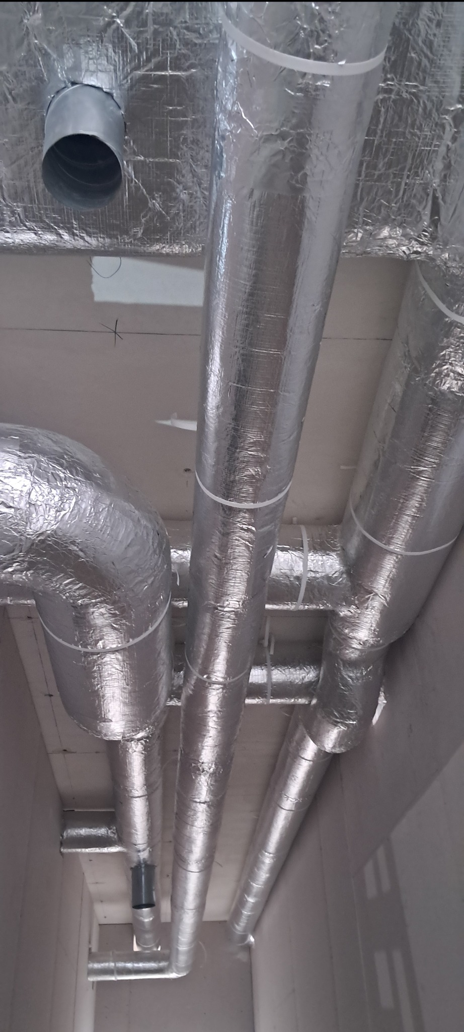 Srebrne, zaizolowane rury wentylacyjne przymocowane do sufitu za pomocą białych opasek zaciskowych, widoczne z perspektywy podłogi w surowym wnętrzu.