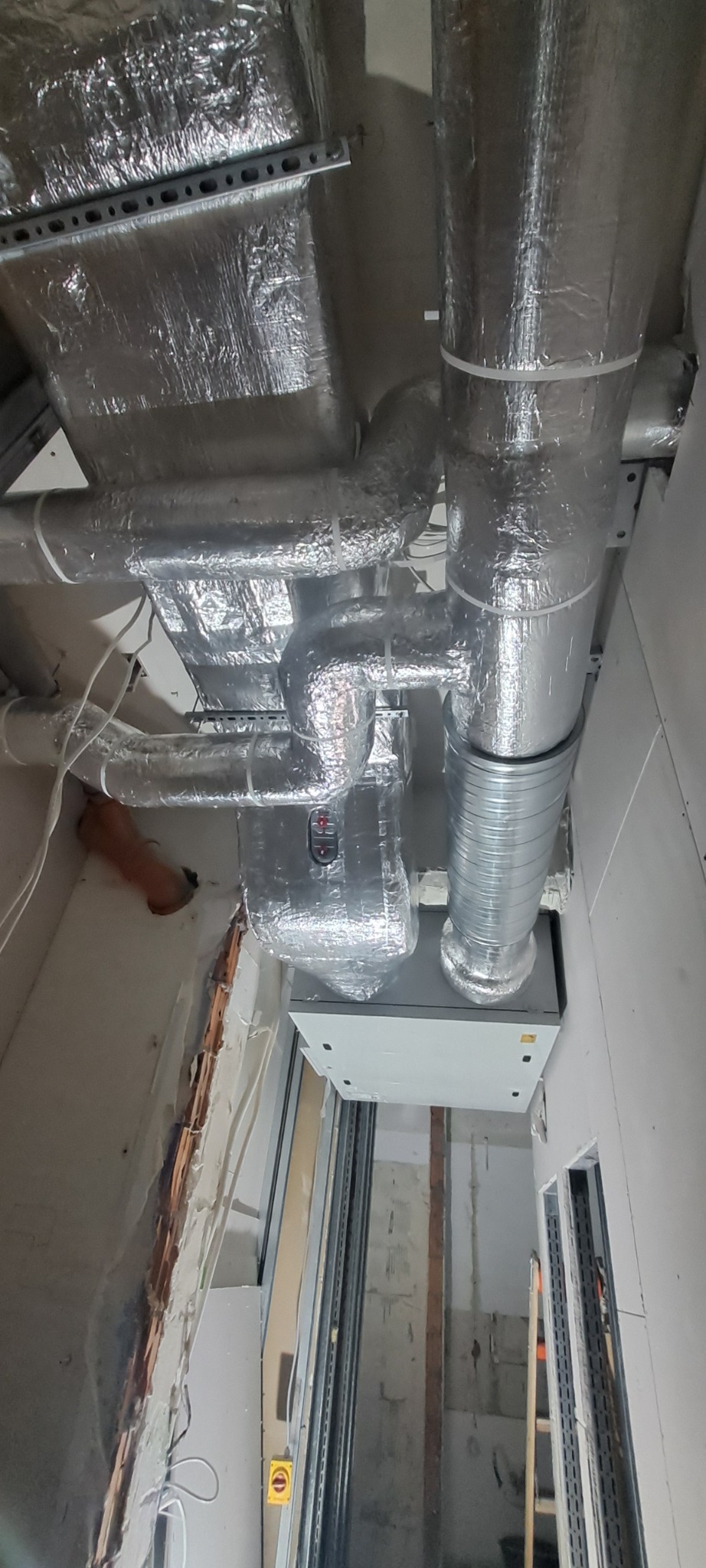 Srebrne, zaizolowane rury wentylacyjne zamontowane pod sufitem w surowym wnętrzu. Widoczne elementy instalacji i konstrukcji wsporczej.