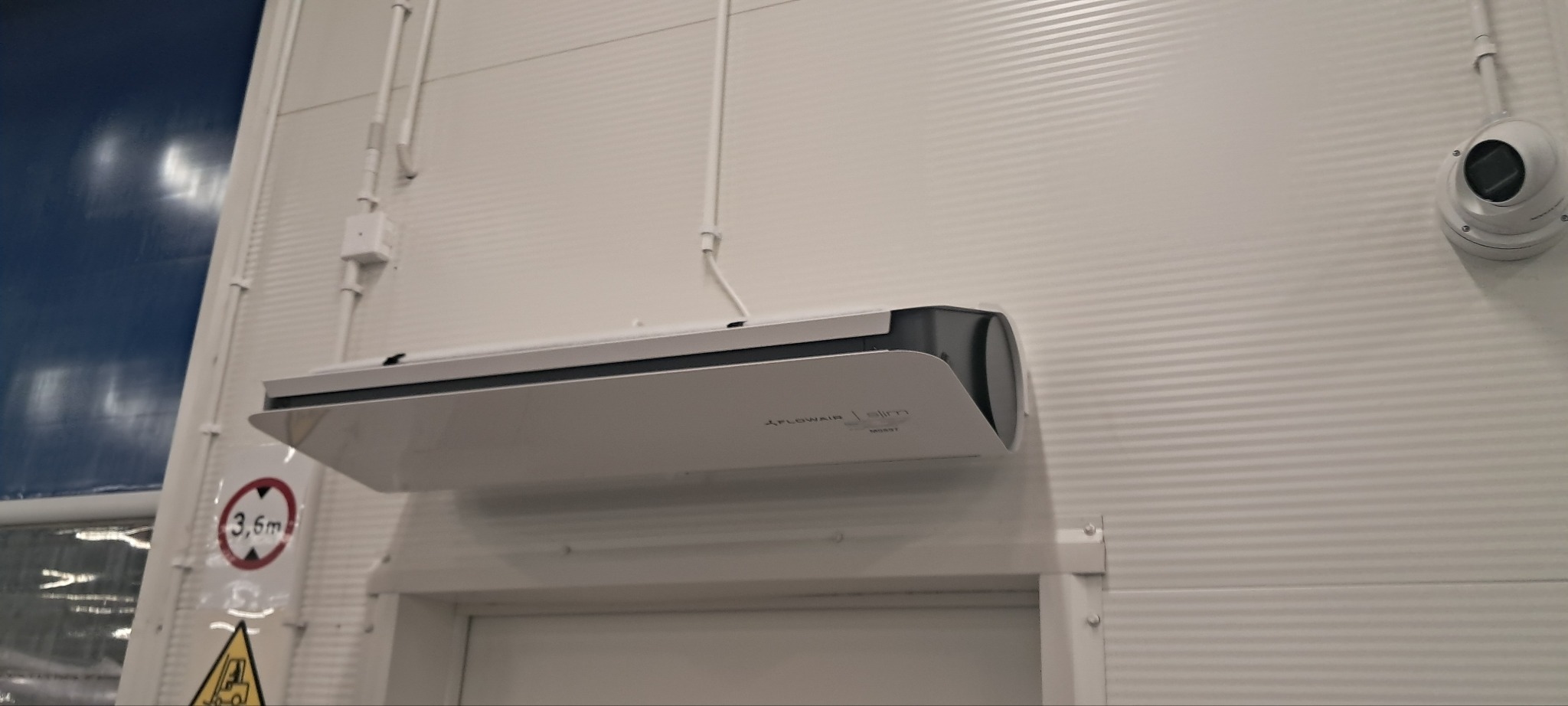 Biała klimatyzacja Flowair Slim zamontowana na ścianie z blachy trapezowej. Widoczna instalacja elektryczna i kamera przemysłowa. Tabliczka informacyjna 3.6m z lewej.