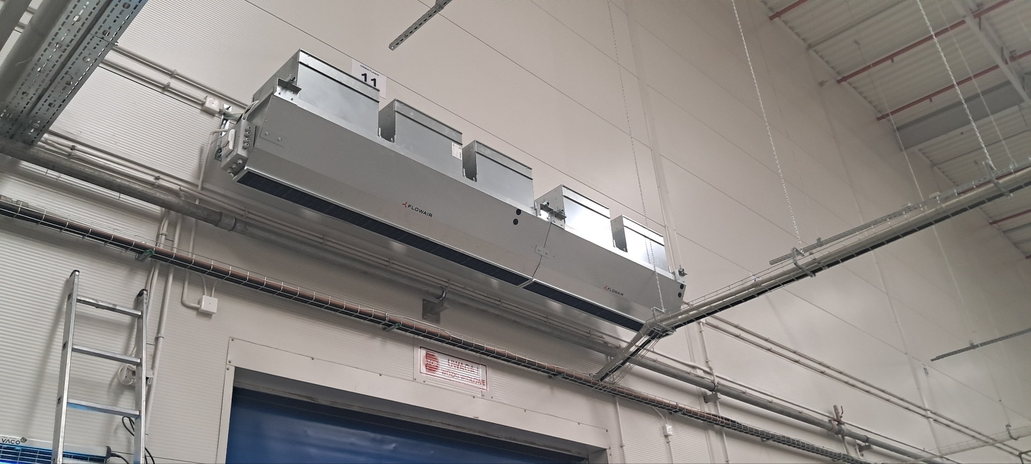 Szara, podwieszona jednostka wentylacyjna Flowair z czterema modułami, zamontowana w hali produkcyjnej. Widoczne rury, kable i drabina.
